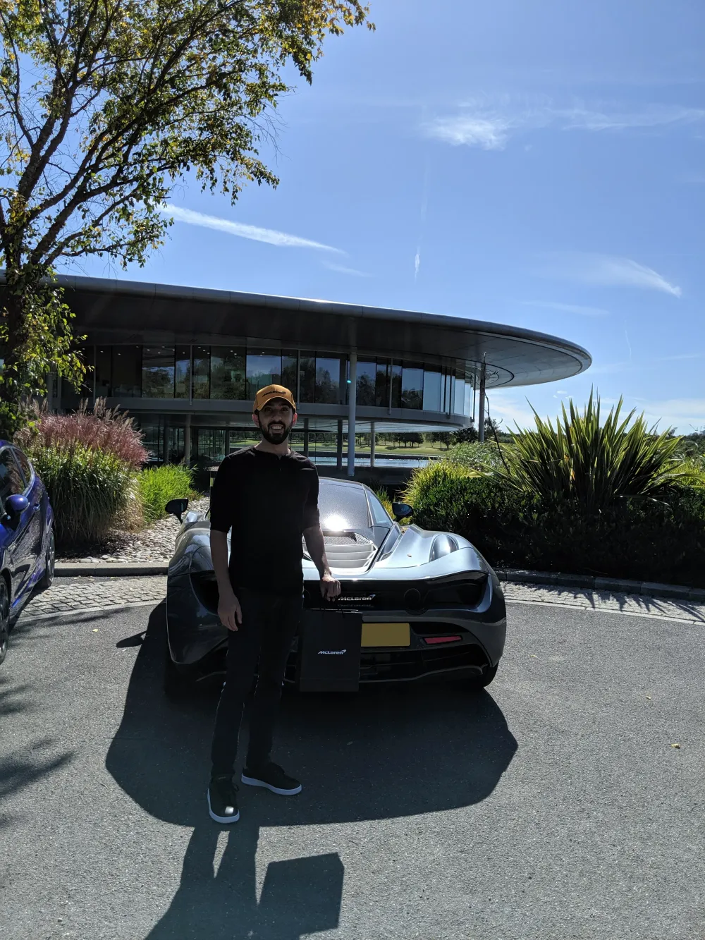 Jimmy Dushku standing outside the McLaren Technology Centre next to a silver McLaren 720S, holding a McLaren shopping bag
